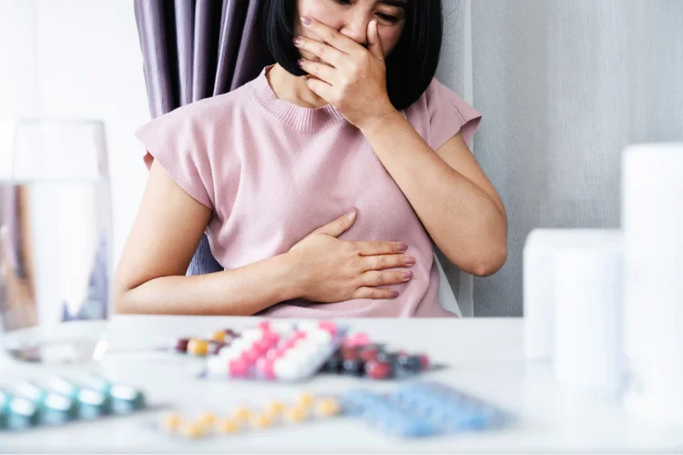 Asian woman feeling nauseous, holding her stomach, with anti-nausea medicine and a glass of water on the table.