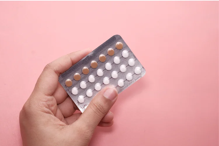 Hand holding combination birth control pill pack with active and placebo pills on pink background.