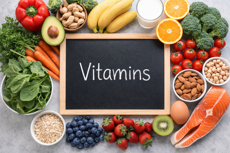 Chalkboard labelled vitamins surrounded by fresh fruits, vegetables, and nuts.