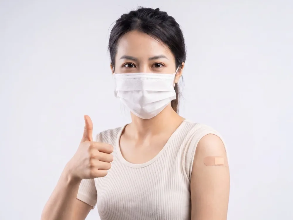 Woman in a face mask and cream top giving a thumbs up with a plaster on her arm