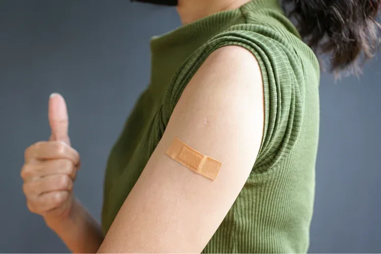 Woman in an olive-green sleeveless top gives a thumbs up, a plaster on her arm