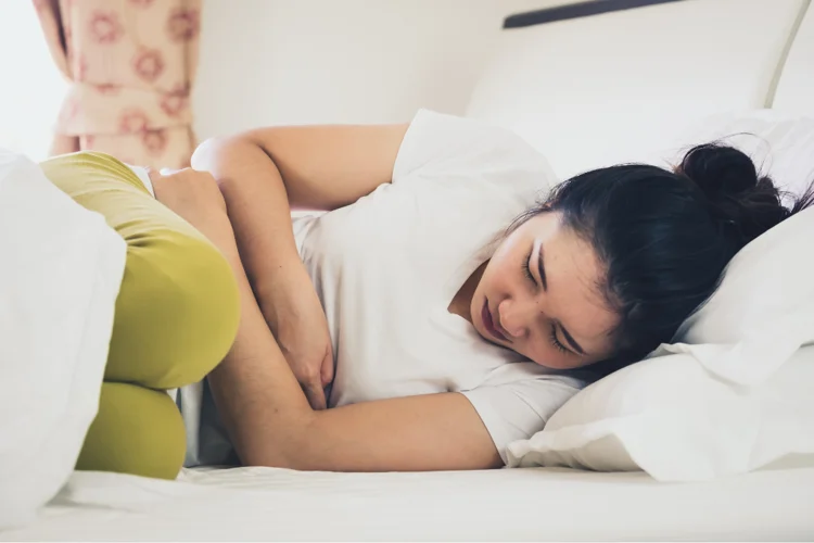 Young Asian woman experiencing nausea, holding her stomach while sitting on a bed at home.