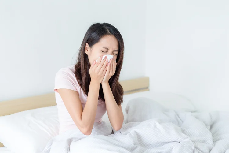 Young woman sneezing into a tissue while sitting in bed, showing flu symptoms such as a runny nose and fatigue.