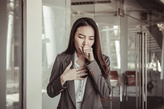 身穿灰色西装外套的女士一手掩口咳嗽，一手按着胸口